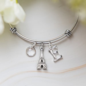 May include: A silver bangle bracelet with three charms: a clear crystal, a miniature Eiffel Tower, and a letter 'E'.