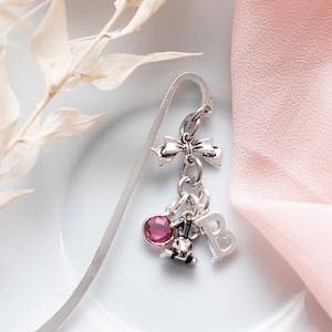 May include: Silver bookmark with a pink crystal charm, a silver crown charm, a silver letter B charm, and a silver bow charm.