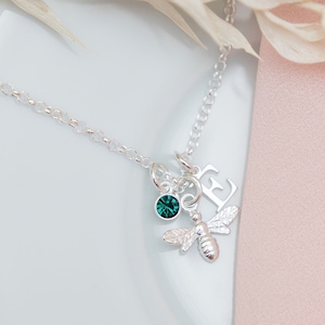 May include: A silver necklace with a bee charm, a small green crystal, and a letter 'E' charm. The necklace is on a white plate with a pink background.