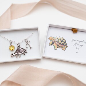 May include: A silver necklace with a turtle charm, a yellow gemstone, and the letter "T" presented in a white gift box. The box also contains a card with a turtle illustration and the text "Your personalised gift message here".