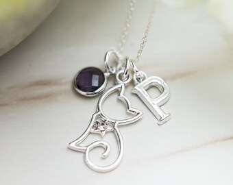 May include: A silver-tone necklace featuring three charms: a circular purple gemstone, a stylized cat outline with a small embedded clear stone, and a capital letter P charm. The charms hang from a delicate silver-tone chain.