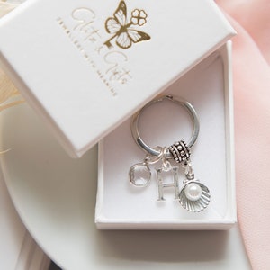 May include: A silver charm keychain with a pearl, a crystal, and a letter H charm. The keychain is in a white box with the logo "Glitter & Gifts Jewellery with Meaning".