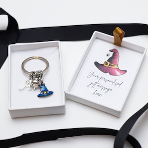 Puede incluir: Un llavero de plata con forma de sombrero de bruja con un dije de sombrero de bruja, una calavera y una letra A. El llavero está en una caja blanca con una tarjeta que dice "Tu mensaje de regalo personalizado aquí".