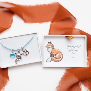 May include: A blue cord necklace with a silver fox charm, a silver initial charm, and a blue gemstone charm. The necklace is in a white box with a card that says "Your personalized gift message here xxx".