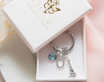 Pode incluir: Um chaveiro de prata com um charme de cristal azul, um charme de letra de prata e um charme da Torre Eiffel de prata. O chaveiro está em uma caixa branca com o texto "Glitter & Gifts Jewellery with Meaning" impresso na tampa.