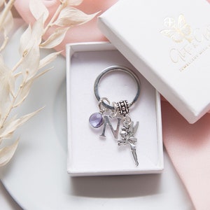 May include: A silver charm keyring with a purple crystal charm, a silver letter N charm, and a silver fairy charm. The keyring is in a white box with the text "Cutie & Co. Jewellery with a Twist" on the lid.
