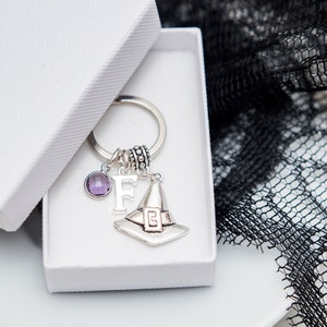 Puede incluir: Un llavero de plata con un dije de sombrero de bruja, una gema púrpura y la letra F. El dije está en una caja de regalo blanca.