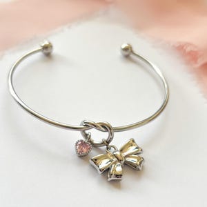 Puede incluir: Pulsera de brazalete plateada con diseño de nudo. Cuenta con un pequeño dije de corazón rosa y un dije de lazo. Una cinta rosa descansa sobre una superficie blanca en el fondo.