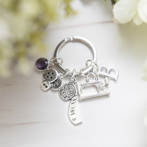 Peut inclure: Porte-clés argenté avec un charme de machine à coudre, un charme de bobine de fil, deux charmes de boutons, un charme de lettre "B" et un charme de pierre précieuse violette.