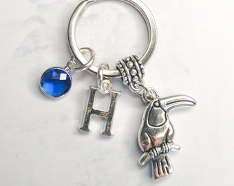 Puede incluir: Un llavero plateado con un dije de cristal azul, un dije de letra H plateada y un dije de tucán.