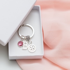 May include: A silver keyring with a pink crystal charm, a silver letter "B" charm, and a silver button charm. The keyring is in a white gift box.