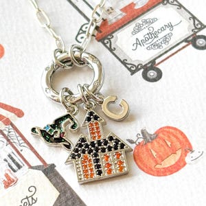 May include: Silver necklace with a charm featuring a Halloween-themed house with orange and black accents, a witch hat, and a letter "G" charm. The background includes a pumpkin illustration and a cart with the word "Sweets".