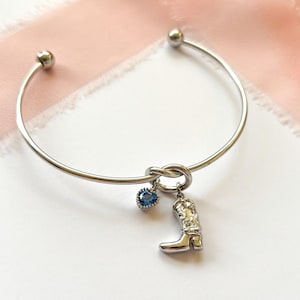 May include: A silver-toned bangle bracelet with a knot design. The bracelet features a small blue gemstone charm and a charm shaped like a cowboy boot. The bracelet is displayed on a white surface with a pink fabric in the background.