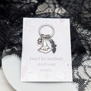 May include: A silver witch hat keychain with a cauldron, bat, and a silver key ring. The keychain is on a white card with the text "Don't let anybody steal your magic."