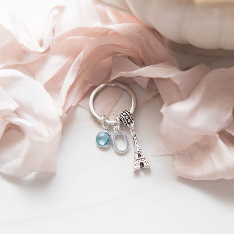 May include: A silver charm bracelet with a blue crystal charm, a silver letter O charm, and a silver Eiffel Tower charm. The bracelet is on a pink fabric background.