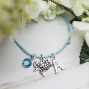 May include: A light blue bracelet with a silver turtle charm, a silver letter A charm, and a blue gemstone charm.