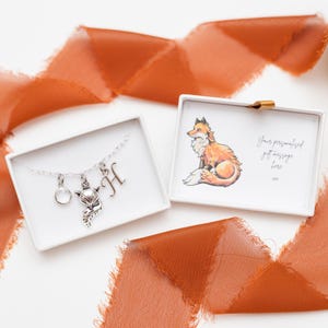 May include: A silver necklace with a fox charm, a circle charm, and an initial charm in a white box. The box has a fox illustration and the text "Your personalized gift message goes here".