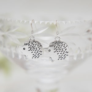 May include: A pair of silver dangle earrings featuring a hedgehog design. The hedgehogs are detailed with small spikes and have a cute, cartoon-like appearance.