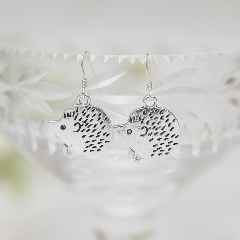 May include: A pair of silver dangle earrings featuring a hedgehog design. The hedgehogs are detailed with small spikes and have a cute, cartoon-like appearance.