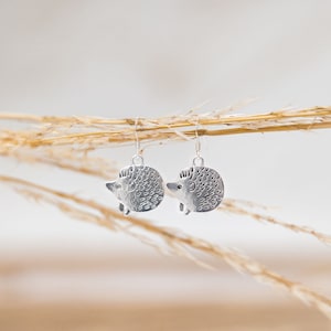 May include: A pair of silver dangle earrings featuring a textured hedgehog design. The earrings are hanging from a branch of dried grass.
