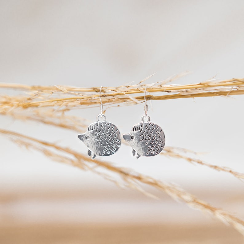 May include: A pair of silver dangle earrings featuring a textured hedgehog design. The earrings are hanging from a branch of dried grass.