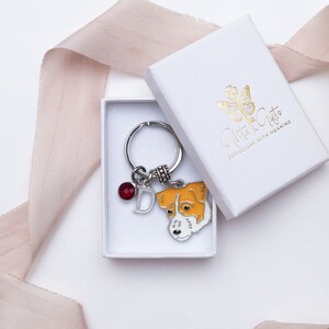 Puede incluir: Una caja de regalo blanca con un logotipo de mariposa dorado y el texto "Gifts & Gifts Joyería con significado". Dentro de la caja hay un llavero plateado con un dije de cristal rojo, un dije de letra plateada "D" y un pequeño dije de perro de esmalte.