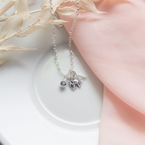May include: A silver necklace with a small elephant charm, a blue gemstone, and the letter "A" hanging from it. The necklace is on a white surface with a pink fabric in the background.