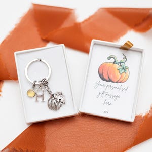 Puede incluir: Una caja de regalo blanca con un llavero de plata con un dije de calabaza en su interior. El llavero tiene una pequeña gema amarilla y un dije con la letra "H". La caja también incluye una tarjeta con una ilustración de acuarela de una calabaza y el texto "Tu mensaje de regalo personalizado aquí xxx".