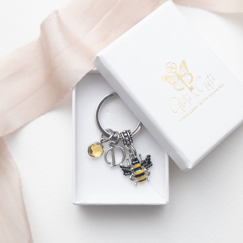 May include: A silver keyring with a bee charm, a yellow gemstone, and the letter "D" charm, presented in a white gift box. The bee charm is black and yellow. The box has gold text and a butterfly logo.