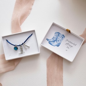 Puede incluir: Un brazalete de cordón azul con un dije de plata de una bota, una piedra preciosa azul y una letra "J". El brazalete está en una caja blanca con una tarjeta que dice "Tu mensaje de regalo personalizado aquí xxx". La tarjeta tiene una ilustración de acuarela de botas de vaquero azules.