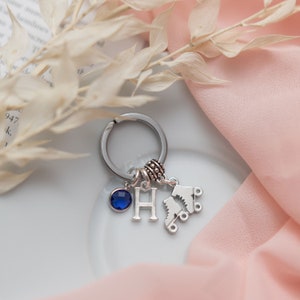 Puede incluir: Llavero de plata con una piedra preciosa azul, un dije de plata con la letra H y un dije de un par de patines.