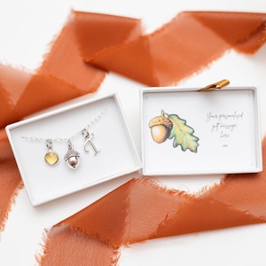 Peut inclure: Un collier en argent avec un pendentif en forme de gland, une petite pierre précieuse et une lettre T. Le collier est dans une boîte blanche avec une petite carte qui dit "Votre message cadeau personnalisé ici".