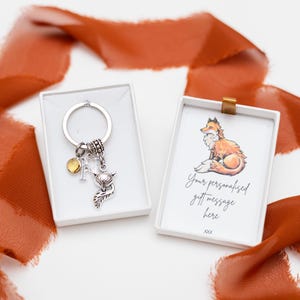 Puede incluir: Un llavero de zorro plateado con un dije y una letra 'N' en una caja blanca. La caja tambi&eacute;n incluye una tarjeta con el texto "Tu mensaje de regalo personalizado aqu&iacute; xxx".