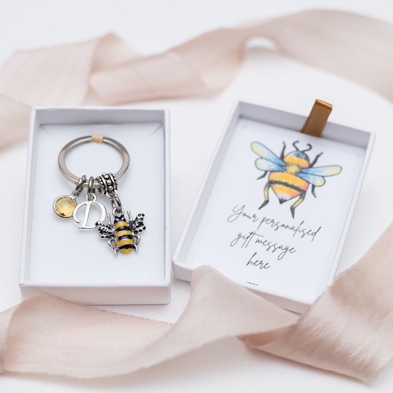 May include: A silver keychain with a bee charm, a yellow gemstone, and a letter "D" charm, presented in a white gift box. The box also contains a card with a watercolor bee illustration and the text "Your personalised gift message here".