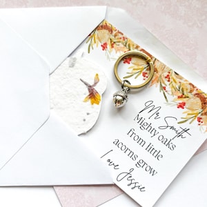 Puede incluir: Un llavero con un dije de bellota plateada y un anillo dorado, sobre una tarjeta con detalles florales y el texto "Mr Smith, Mighty oaks from little acorns grow, I love Jessie." La tarjeta está dentro de un sobre blanco.