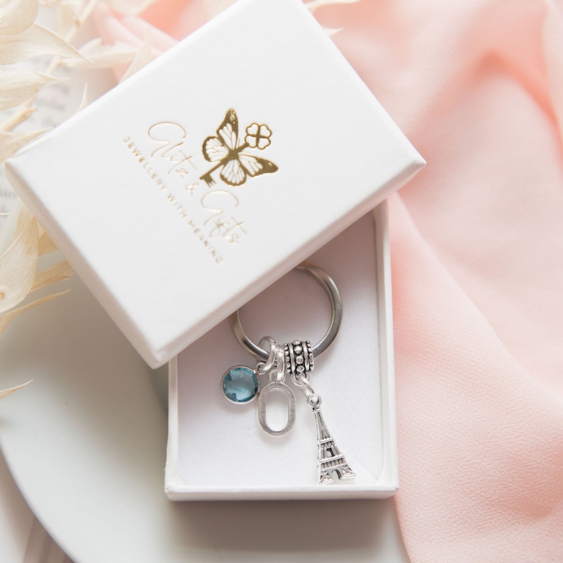 May include: A silver charm keychain with a blue crystal charm, a silver letter charm, and a silver Eiffel Tower charm. The keychain is in a white box with the text "Glitter & Gifts Jewellery with Meaning" printed on the lid.