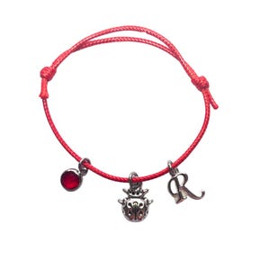May include: Red string bracelet with a ladybug charm, a red gemstone charm, and a silver charm with the letter "R".