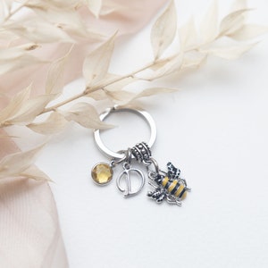 May include: Silver keychain with a citrine gemstone, a letter "D" charm, and a bee charm with black and yellow stripes. The keychain is a personalized accessory.