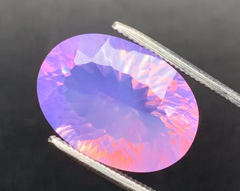 May include: An oval-shaped gemstone with a faceted surface, held by silver tweezers. The gem displays a gradient of colors, transitioning from pink to purple, with hints of orange. The background is a dark gray.