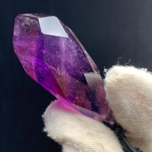 May include: A vibrant purple amethyst crystal with a faceted, pointed shape. The crystal displays a gradient of color, transitioning from deep purple to lighter hues. The crystal is held by a white glove against a dark background.