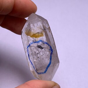 May include: A clear quartz crystal with a double termination, also known as a double-pointed crystal. The crystal has a natural inclusion of a dark grey mineral and a yellow mineral. The crystal has a blue ink circle drawn on it.