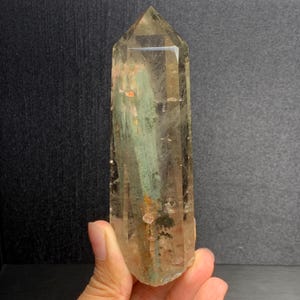 May include: A clear, hexagonal quartz crystal point featuring internal inclusions of green chlorite and small orange mineral deposits. The crystal has a polished, faceted termination and a translucent body that reveals natural internal fractures.