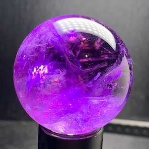 Könnte beinhalten: Eine polierte, kugelförmige Amethyst-Kristallkugel. Die Kugel hat eine tiefe lila Farbe mit inneren kristallinen Strukturen und Einschlüssen. Die Oberfläche reflektiert das Licht und erzeugt helle Glanzlichter. Die Kristallkugel steht auf einem schwarzen Sockel, vor dunklem Hintergrund.