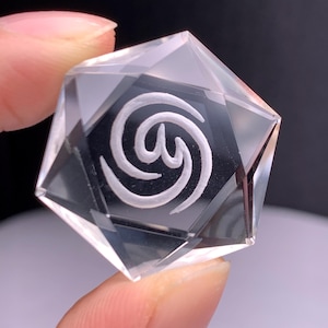 Puede incluir: Un cristal transparente y facetado con un diseño de espiral blanco grabado sobre un fondo negro. El cristal se sostiene entre los dedos, mostrando su forma geométrica y sus intrincados detalles. El cristal es un objeto decorativo.
