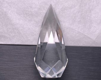 May include: A clear, faceted crystal teardrop pendant. The pendant has a pointed top and a geometric design. The crystal is transparent, allowing light to pass through. The pendant is displayed on a dark surface.