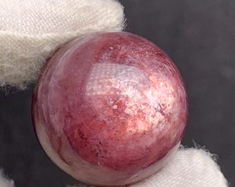 May include: A polished, spherical gemstone with a reddish-pink hue. The surface has a mottled appearance with variations in color and texture. The gem is held by a white fabric.