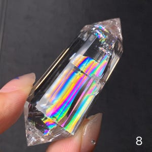May include: A clear quartz crystal with a pointed tip. The crystal refracts light, displaying a spectrum of rainbow colours. The crystal is held in a hand against a dark background. The number "8" is in the lower right corner.