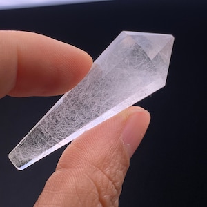 May include: A clear quartz crystal point with a rough, textured surface. The crystal is shaped like a long, thin point with a rounded tip.