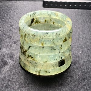 Puede incluir: Una pila de cinco pulseras de piedras preciosas translúcidas de color verde claro. Las pulseras tienen un diseño circular con inclusiones de color verde oscuro y negro. Las pulseras están apiladas, creando una torre de joyería de piedras preciosas.