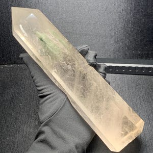May include: A large, elongated, clear quartz crystal point with a hexagonal shape. The crystal is transparent with visible internal fractures and inclusions, featuring a pointed termination at one end and a flat base at the other end.